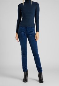 Haut à col roulé côtelé bleu marine, associé à un pantalon en velours côtelé bleu foncé avec une coupe ajustée et des poches latérales, complété par des bottines noires.