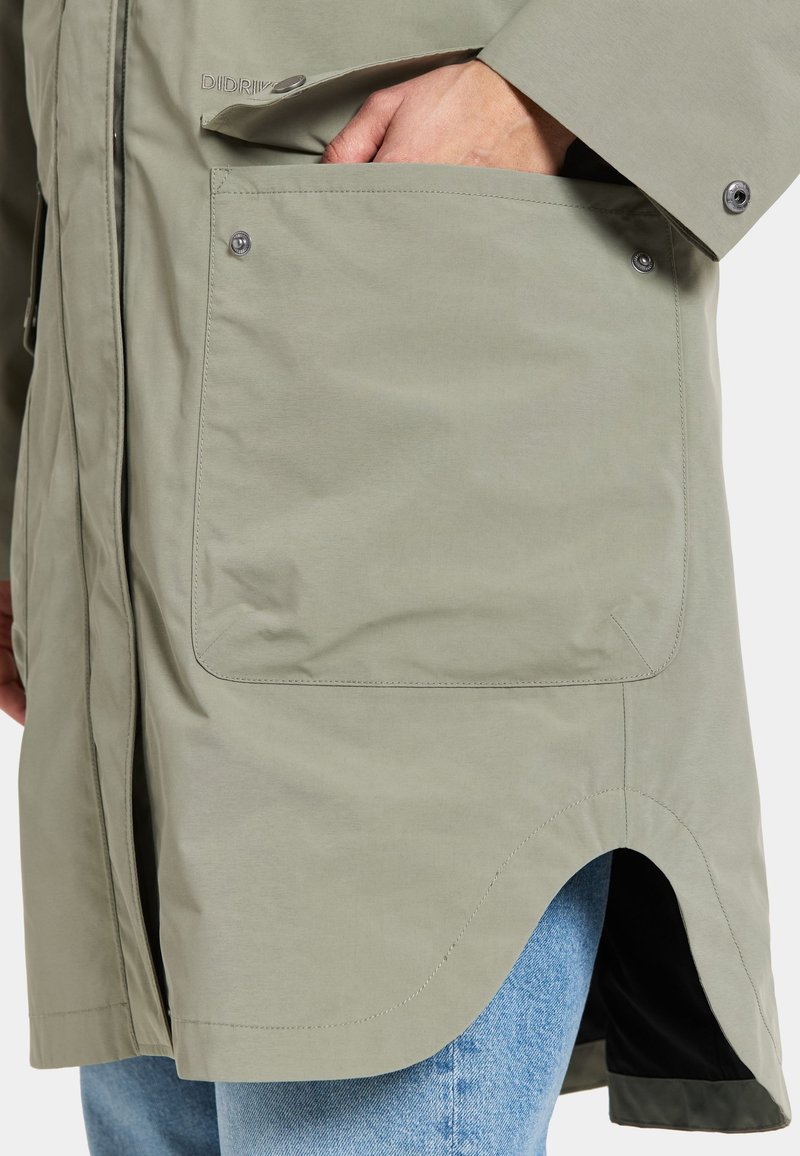 Helle olivgrüne Jacke mit einer großen, flachen Tasche mit Druckknopf, abgerundetem Saum und seitlichen Schlitzen. Hergestellt aus glattem, wasserabweisendem Material.