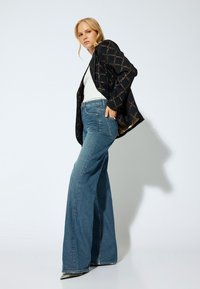 Blazer negro con estampado, blusa blanca y jeans acampanados de un azul claro. Tejido texturizado, diseño de pierna ancha, tacones plateados puntiagudos añaden detalle.
