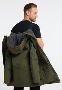 Olivgrön parkas med huva, med dragkedjestängning, ribbad innerfoder och sidofickor. Materialet verkar vara vattentåligt med en slät textur.
