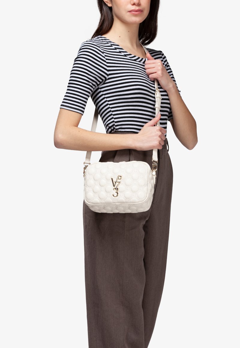 Borsa a tracolla bianca trapuntata con motivi circolari, hardware dorato e tracolla rimovibile, indossata sulla spalla sopra una maglietta a righe.