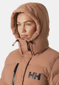 Chaqueta de plumas marrón claro con capucha alta y ajustable, que presenta un cierre frontal con cremallera, un logo negro en el pecho y un tejido texturizado.