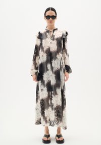 Vestido maxi fluido y plisado en colores claros con patrones florales. Presenta un escote con lazo y mangas largas. Combinado con sandalias negras y gafas de sol.