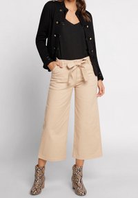 Zwarte jas met gouden knopen over een geribbelde zwarte top, gecombineerd met beige wijde broek en slangenprint enkellaarsjes.
