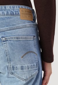 Vaqueros de G-Star RAW en azul claro con un parche de cuero en la cinturilla. Las características incluyen un bolsillo trasero con costuras amarillas y un diseño curvado.