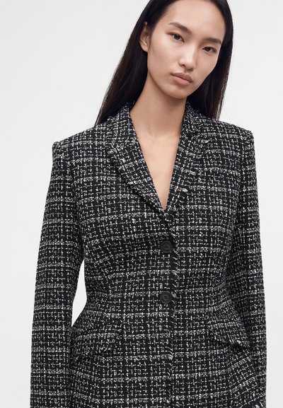 Femme aux longs cheveux foncés portant un blazer en tweed noir et blanc avec deux boutons noirs et des revers crantés.