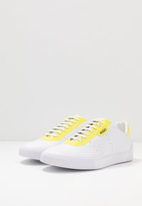 Scarpe da ginnastica bianche con accenti gialli, pannelli con lacci testurizzati e suola in gomma. Presentano un logo sul lato e un design minimalista.