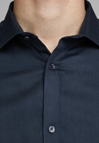 Närbild av en mans hals och en mörk texturerad skjorta med knappar, där två knappar är knäppta och kragen är öppen.