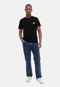 Schwarzes T-Shirt mit einem kleinen gelben Zitronenmotiv auf der linken Brust, kombiniert mit blauen Jeans und weißen Sneakern. Lässiger Schnitt und schlichte Gestaltung.