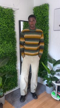 Sweater riscado em verde, castanho e bege; calças cargos de cor clara com remendos; botas pretas; fundo com vegetação e um espelho.