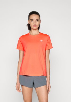 Femme portant une t-shirt sportif à manches courtes New Balance orange vif et un short de course gris, se tenant contre un fond blanc uni.
