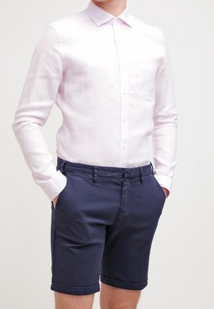 Mand iført en langærmet hvid skjorte med knapper, stoppet ned i mørkeblå skræddersyede shorts, med hænderne i lommerne.