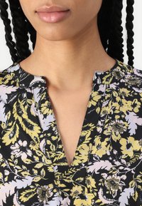 Blomstret bluse med sort bakgrunn, med gule og lavendelblomster mønstre. V-hals design med teksturert finish og liten krage.