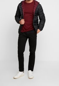 Schwarze Daunenjacke mit burgunderfarbener Innenauskleidung, getragen über einem burgunderfarbenen Pullover, kombiniert mit schwarzen Hosen und weißen Sneakern. Glattes Material, Reißverschlussdetails.