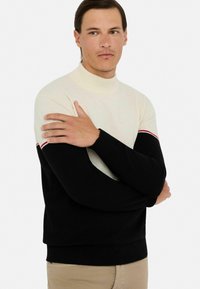 Turtleneck-Pullover mit einem cremefarbenen oberen Teil und einem schwarzen unteren Teil mit einem roten Akzentstreifen. Gestrickte Textur und gerippte Bündchen.