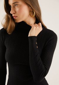 Svart ribbestrikket turtleneck genser med teksturert mønster og tettsittende design. Har knappdetaljer på ermene. Mykt strikket materiale.