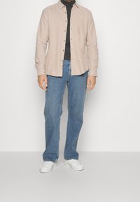 Jasnobeżowa koszula na guziki na czarnym golfie, zestawiona z luźnymi niebieskimi dżinsami i białymi trampkami. Prosty, casualowy styl.