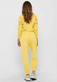 Chándal de algodón amarillo con un ajuste holgado, que presenta un cuello redondo acanalado, puños y cinturilla elásticos. Combinado con zapatillas deportivas grises.