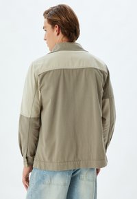 Beige en olijfgroene jas met een kraag, lange mouwen en knoopsluitingen aan de manchetten. Voorzien van contrasterende panelen en gestikte details. Casual ontwerp.