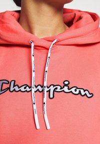Sweat à capuche corail avec cordons blancs présentant un logo répété. Tissu doux, style pullover, et nom de la marque brodé en noir sur le devant.