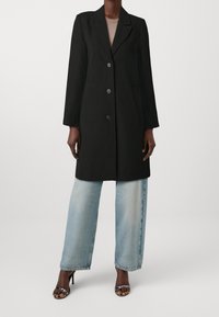 Abrigo negro a medida con solapa con muesca, tres botones y bolsillos laterales, combinado con jeans de pierna ancha y lavado claro y tacones negros.