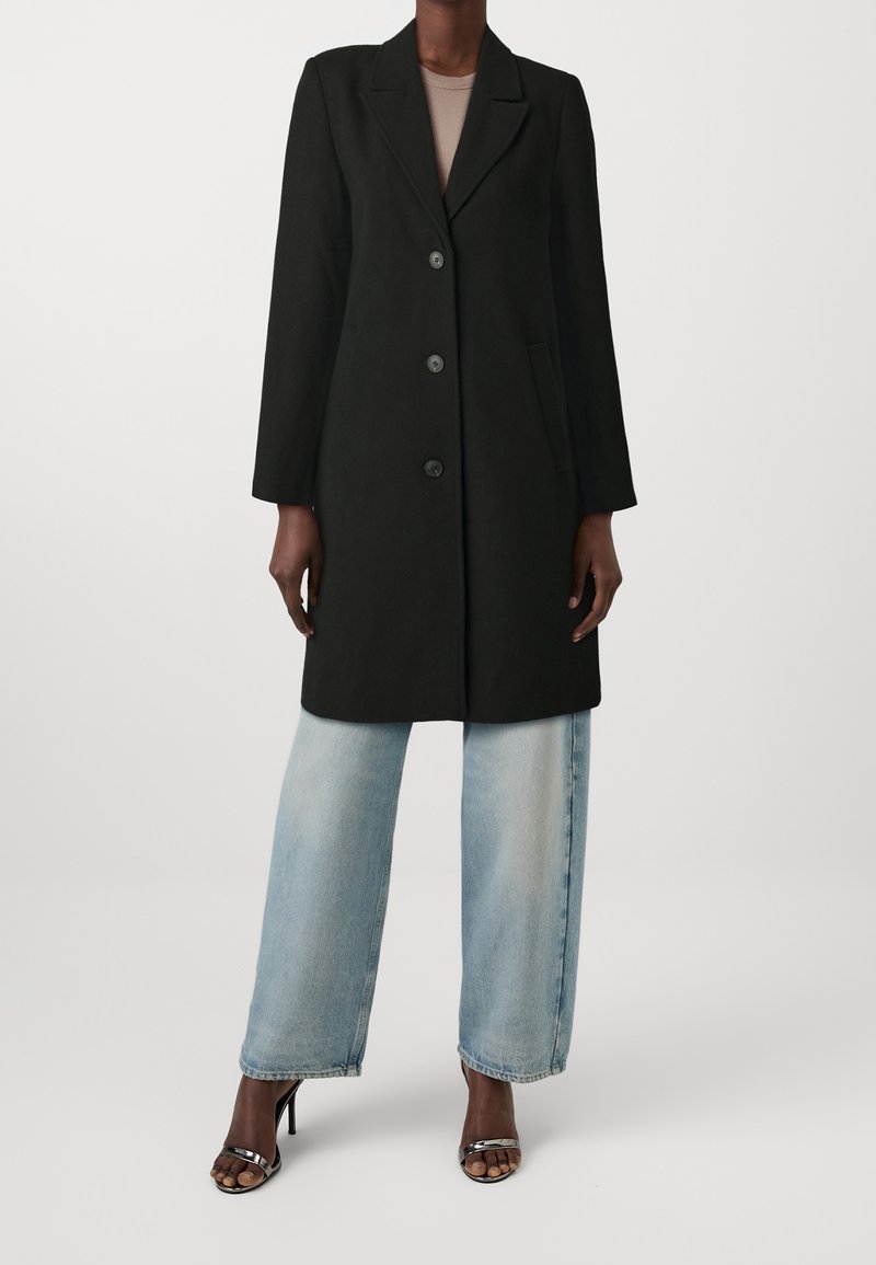 Abrigo negro a medida con solapa con muesca, tres botones y bolsillos laterales, combinado con jeans de pierna ancha y lavado claro y tacones negros.