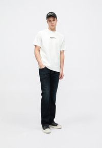 T-shirt de algodão branca com logótipo preto, combinada com jeans azul escuro. Ténis com parte superior em tela preta e sola de borracha branca.