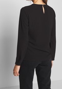 Top nero a maniche lunghe con collo rotondo e dettaglio a chiave sul retro. Tessuto morbido e vestibilità comoda, abbinato a jeans scuri.