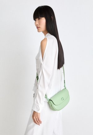 Femme aux longs cheveux noirs portant une robe blanche avec des découpes aux épaules et un sac bandoulière vert clair sur le côté sur fond blanc.