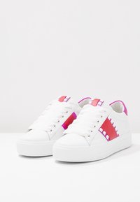 Baskets blanches avec une texture lisse, présentant des accents rose vif et rouge sur les côtés, et des clous carrés pour un détail supplémentaire.
