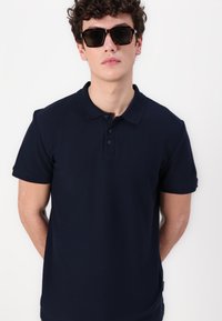 Námořnická modrá polo košile z texturované látky, s klasickým límcem a třemi knoflíky, doplněná tmavými čtvercovými slunečními brýlemi.