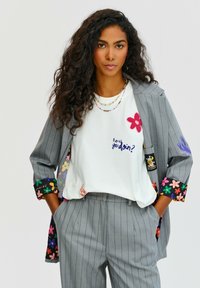 Šedý pruhovaný oblek s bílým tričkem, které má barevnou květinovou výšivku a text. Sako má rukávy s květinovým vzorem. Texturované látky.