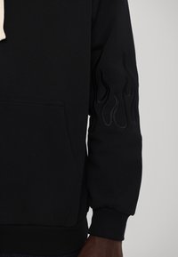 Sudadera negra con bolsillo frontal y un sutil diseño de llamas grabado en la manga derecha, usada por una persona de piel oscura.