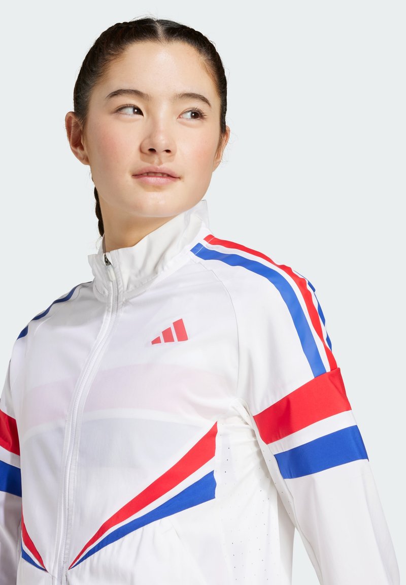 Chaqueta deportiva blanca con rayas rojas, azules y blancas. Presenta un collar alto, cierre con cremallera y secciones perforadas para ventilación.