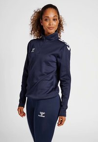 Haut de sport bleu marine à fermeture éclair avec logo blanc et accents aux épaules, assorti à un legging coordonné. Texture lisse, coupe ajustée.
