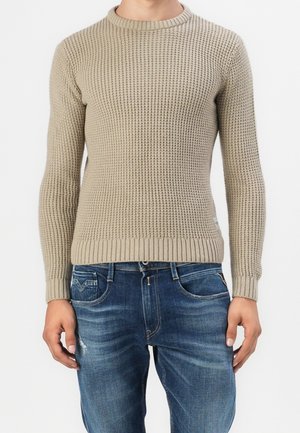 Beige strukturierter Strickpullover, getragen mit verblichenen blauen Jeans, bei einer Person, von Hals bis Hüfte vor einem weißen Hintergrund abgebildet.