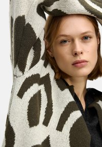 Knit sweater featuring brown and cream abstract patterns, round neckline, and soft texture. Model poses with one arm raised.