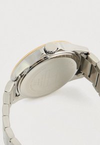 Hõbedane randmekell metallist linkkete ja ümmarguse kuldse raami ning reguleerimisnupuga, nähtud tagantpoolt valgel taustal.