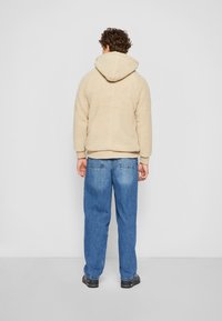 Hoodie en polaire beige avec capuche, présentant une surface texturée. Jean en denim bleu ample avec poches arrière. Chaussures noires.