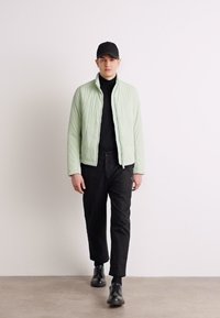 Jachetă pufosă verde deschis, pulover negru cu guler, pantaloni tapered negri, șapcă neagră și pantofi negri, pe un fundal neutru.