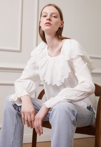 Blusa de algodão branca com decote e mangas rendadas, apresentando bordado de ilhós. Combinada com jeans de cintura alta azuis e sentada numa cadeira de madeira.