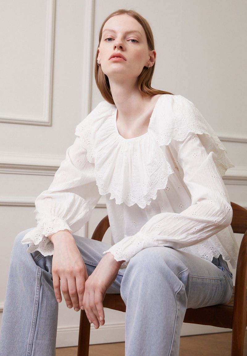 Blusa de algodão branca com decote e mangas rendadas, apresentando bordado de ilhós. Combinada com jeans de cintura alta azuis e sentada numa cadeira de madeira.