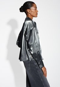 Metaalachtige zilveren overhemd met een losse pasvorm, lange mouwen en een kraag, in combinatie met donkere denim jeans, getoond vanuit een zijprofiel.