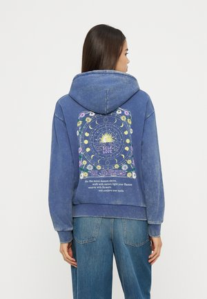 Naine, kes kannab sinist kapuutsi, millel on päike, kuu faasid, lilled ja tekst isearmastusest ning õnnistustest seljal, kombineeritud siniste teksadega.