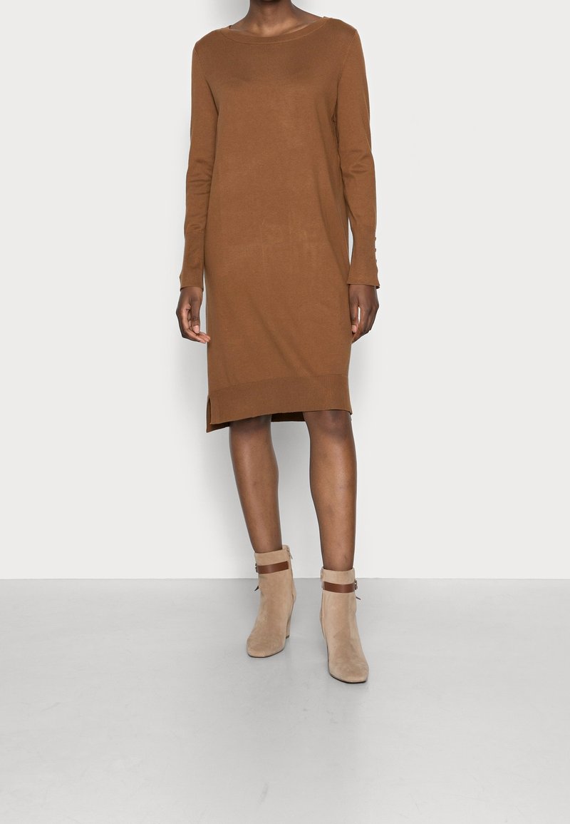 Personne portant une robe marron à manches longues longueur genou et des bottines beige avec des sangles marron, debout sur un fond uni.