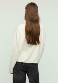 Cremfarbener Pullover mit lockerer Passform, gerippten Bündchen und Saum. Kombiniert mit hochgeschnittenen schwarzen Jeans. Die Haare sind lang und wellig. Der Hintergrund ist schlicht.