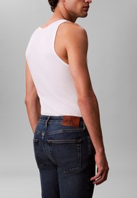 Mand iført en hvid ærmeløs skjorte og mørkeblå jeans med en brun Calvin Klein læderlap på linningen, stående mod en grå baggrund.