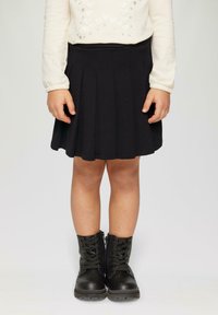 Jupe plissée noire, longueur genou, fabriquée en matériau doux. Associée à des bottes noires à lacets avec une finition texturée et une fermeture éclair sur le côté.