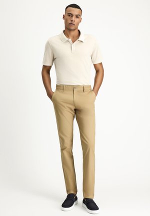 Beige polo shirt and tan slim-fit trousers with a smooth texture, paired with navy slip-on shoes and a simple button detail on the collar.