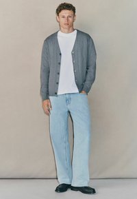 Tânăr îmbrăcat cu un cardigan gri cu nasturi, tricou alb, blugi deschiși la culoare și pantofi negri, stând pe fondul unei suprafețe bej simple.
